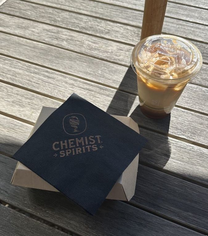 Iced coffee in a clear cup with ice on a sunlit wooden patio, beside a dark napkin that reads 'Chemist Spirits' placed on a small cardboard box.