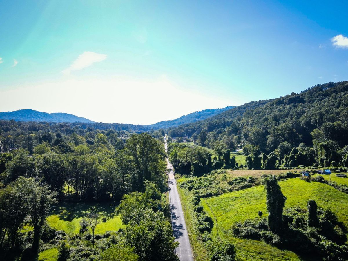 Aerial view of the Ecusta Trail corridor between Hendersonville and Horse Shoe, showcasing green mountain landscapes, open fields, and a rural country road on a sunny day.