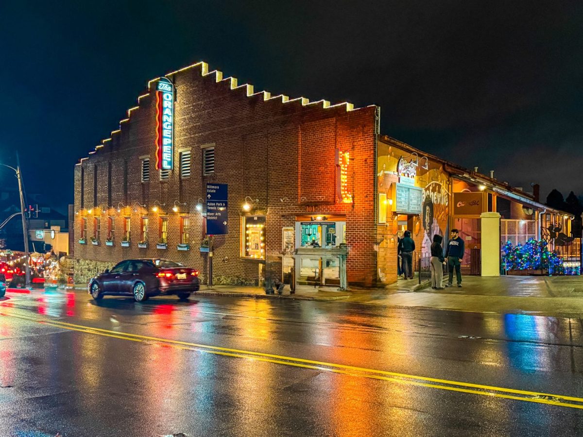 The Orange Peel music venue illuminated at night in downtown Asheville, NC, with glowing marquee lights, colorful reflections on rain-soaked streets, and visitors gathering outside before a live show.
