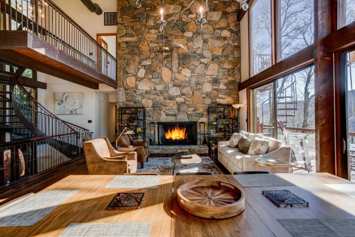 Spacious mountain-view living room at a luxury Asheville vacation rental, featuring a floor-to-ceiling stone fireplace, modern rustic décor, large picture windows with Blue Ridge Mountain views, and cozy seating ideal for relaxing getaway stays.