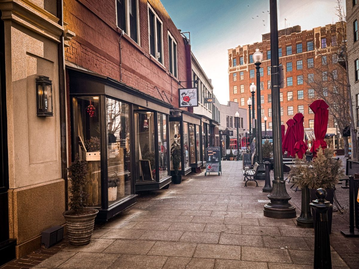 Shops and boutiques along Wall Street in downtown Asheville, NC, with brick storefronts, sidewalk seating, red patio umbrellas, and historic buildings lining the pedestrian-friendly street.