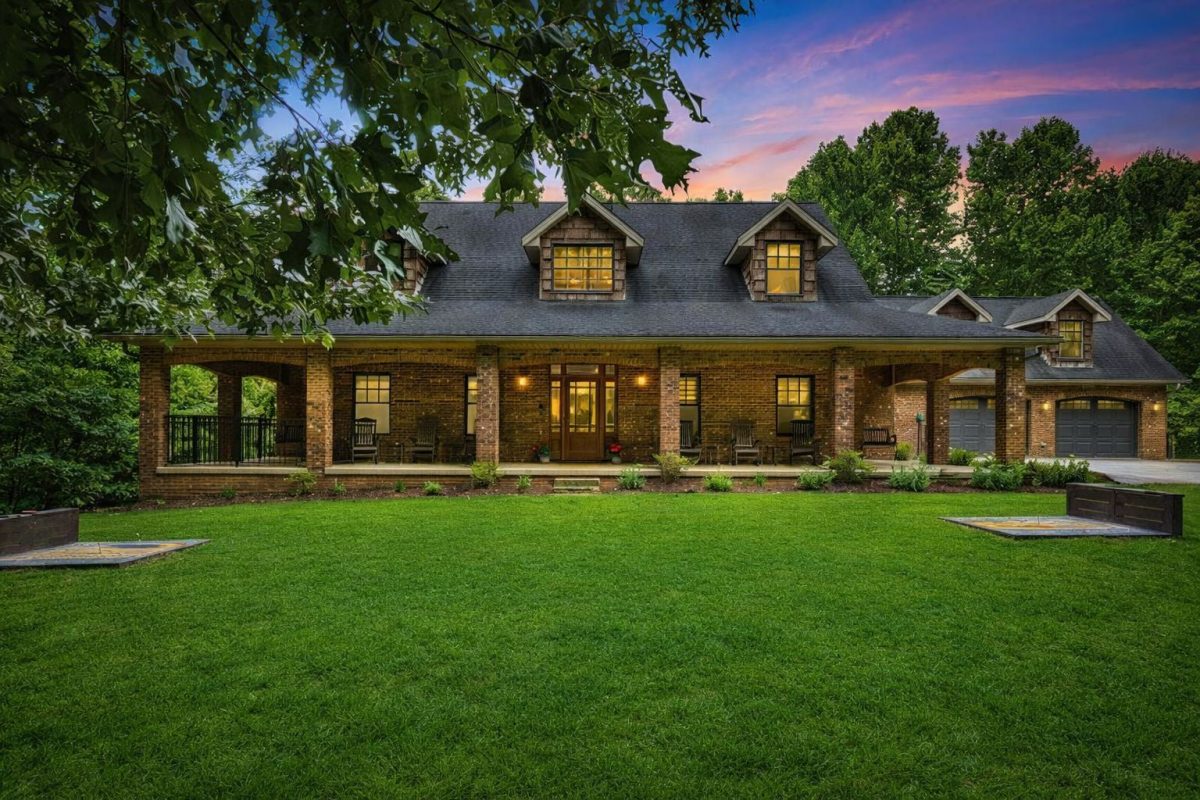 Large group vacation rental in Western North Carolina with a spacious green lawn, wraparound porch, and mountain setting at sunset.