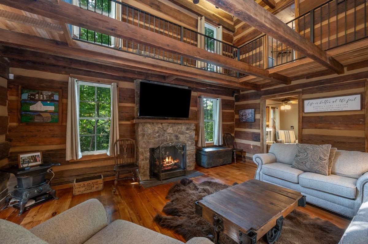 Rustic interior living room at Russell’s Fort in Lake Lure, North Carolina, featuring exposed log beams, a stone fireplace, loft balcony, and cozy mountain lodge furnishings.