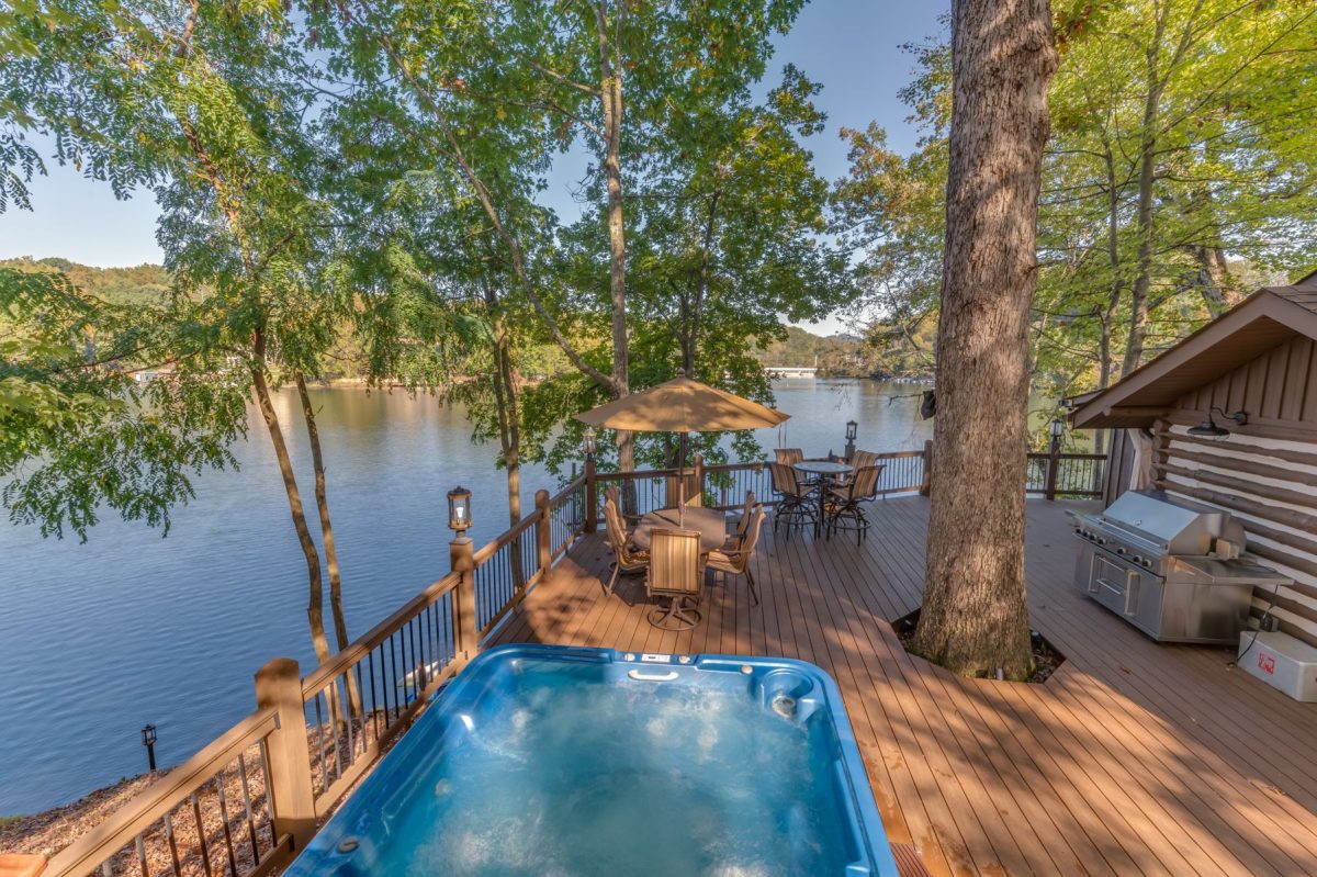 Lakefront log cabin deck in Lake Lure, North Carolina featuring a private hot tub, outdoor dining area, grill, and peaceful water views surrounded by trees.