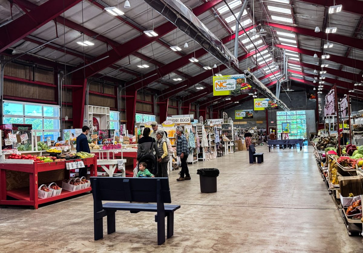 Visitors explore the spacious indoor vendor hall at the WNC Farmers Market in Asheville, NC, featuring fresh produce, local crafts, farm goods, and regional products beneath exposed red steel beams.