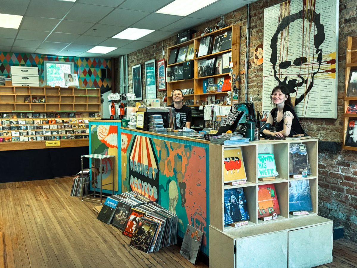 Friendly staff members behind the counter at Harvest Records in West Asheville, surrounded by vinyl albums, colorful artwork, exposed brick walls, and independent music shop decor.