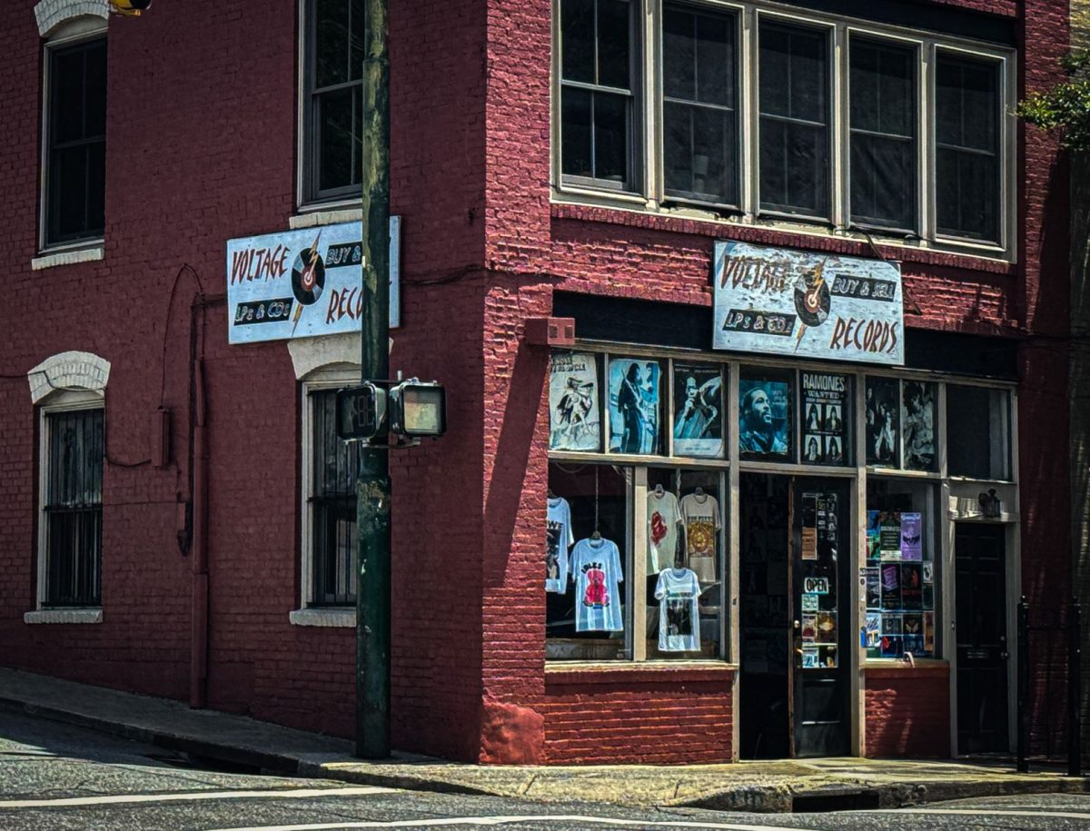 Exterior corner view of Voltage Records in downtown Asheville, featuring a vintage brick storefront, classic record shop signage, music posters, and band merchandise displayed in the windows.