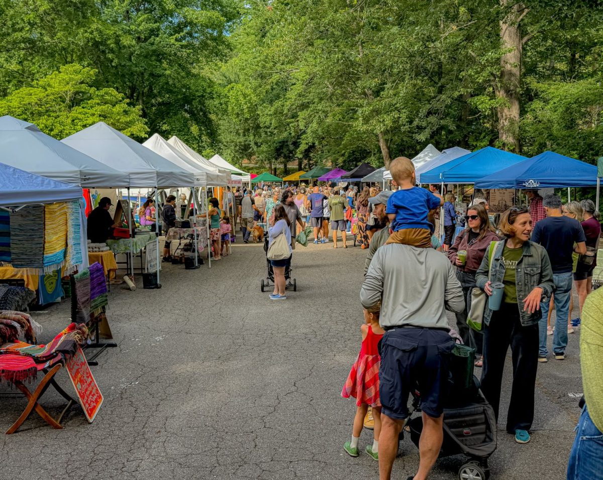 Crowds of shoppers browse vendor tents at the North Asheville Tailgate Market, exploring local produce, artisan goods, handmade crafts, and fresh foods beneath a canopy of mature trees.