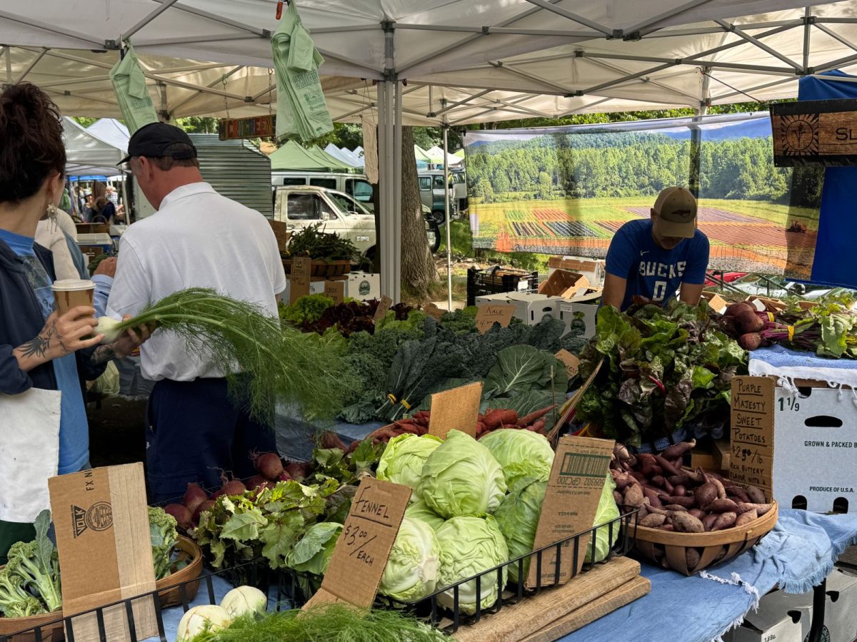 Fresh vegetables including cabbage, kale, beets, sweet potatoes, and fennel are displayed at a local farm stand inside the North Asheville Tailgate Market as shoppers browse seasonal produce.