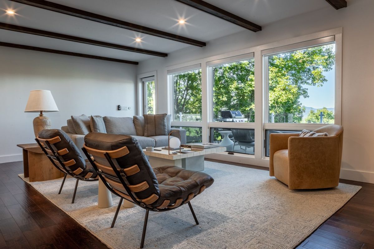 Bright and modern living room at Sunset Sanctuary featuring designer lounge chairs, a plush sofa, floor-to-ceiling windows, and access to a mountain-view deck surrounded by trees.