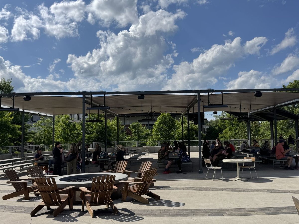 Guests gather on the expansive outdoor patio at New Belgium Brewing in Asheville, featuring shaded seating areas, a central fire pit, and views of the surrounding greenery under a bright summer sky.