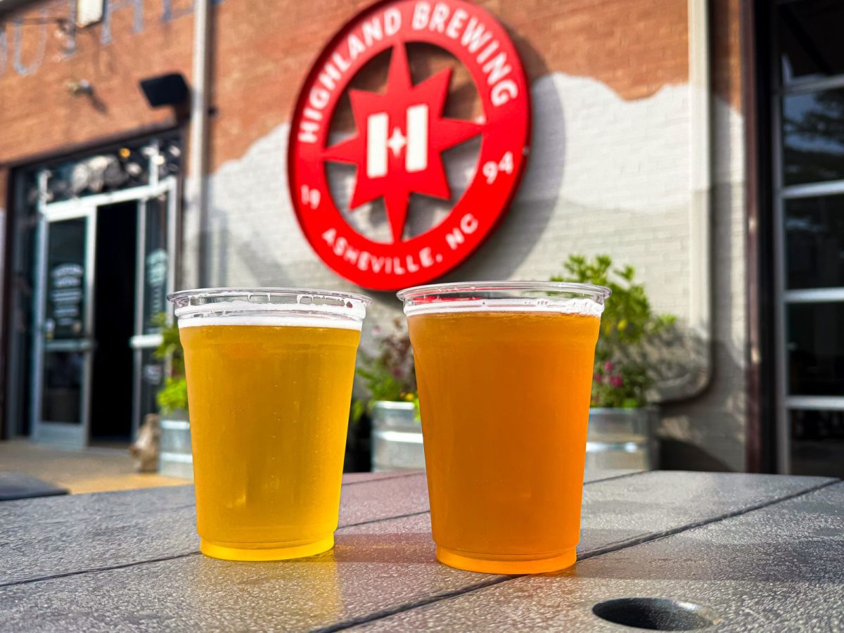 Two craft beers on an outdoor table at Highland Brewing Company in Asheville, North Carolina, with the brewery’s iconic red logo displayed in the background.