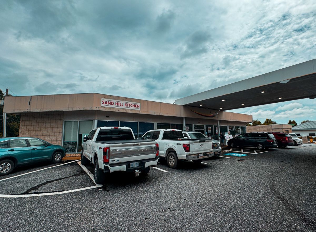 Exterior view of Sand Hill Kitchen in Enka, North Carolina, showing the restaurant's storefront and parking area within a working gas station complex beneath dramatic mountain skies.