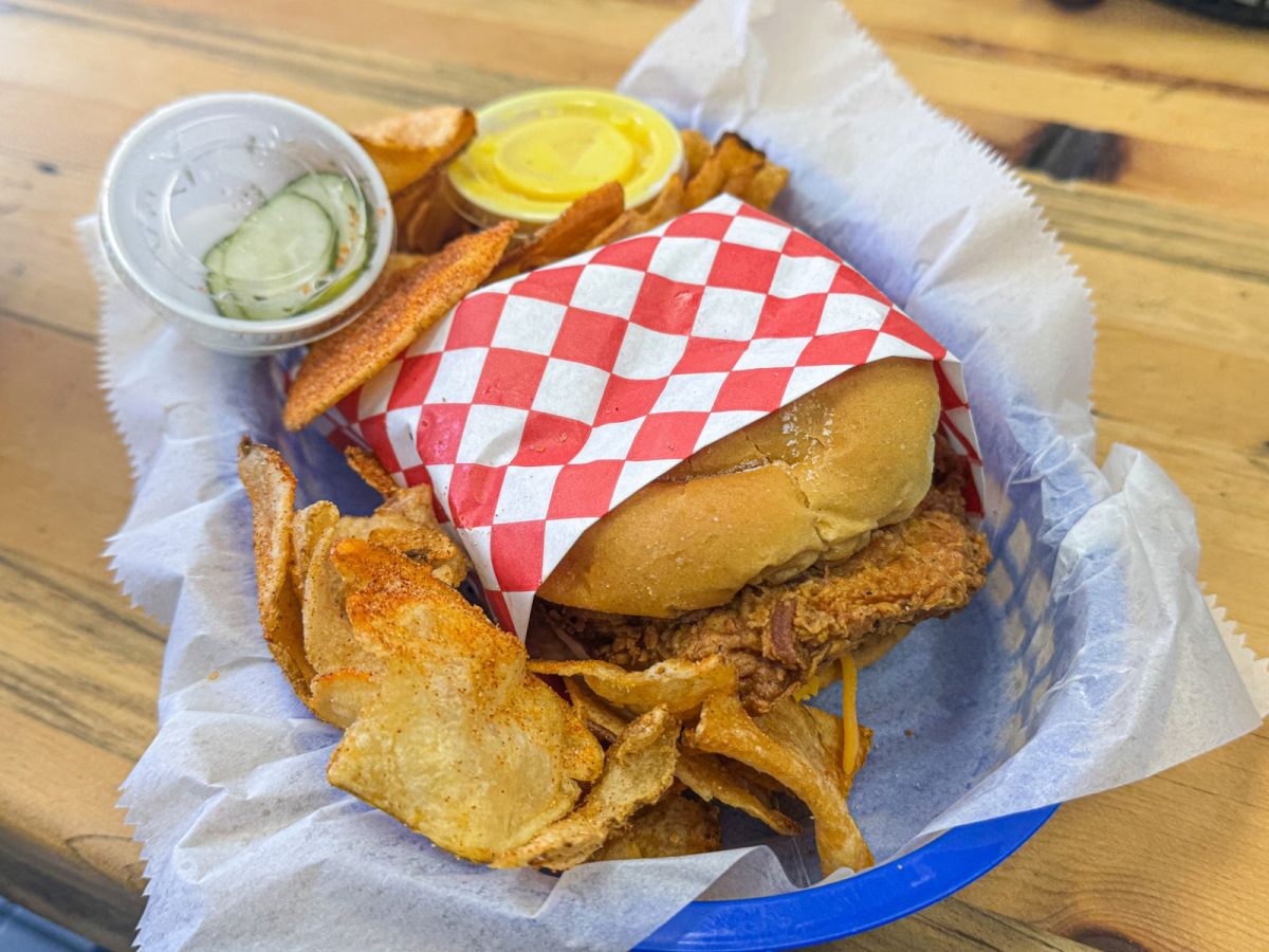 Close-up of Sand Hill Kitchen's fried chicken sandwich served with house-made potato chips, pickle slices, and honey-mustard sauce on a wooden table in Enka, North Carolina.