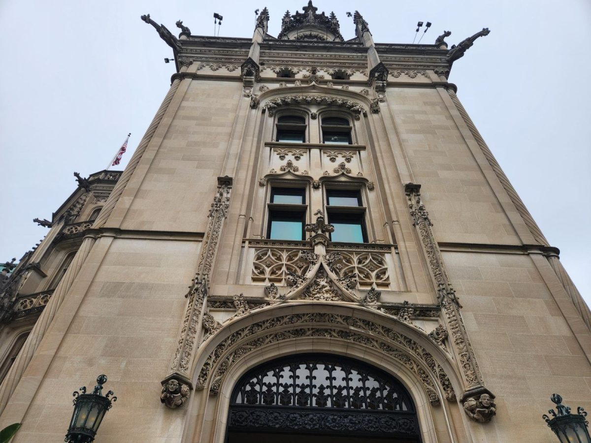 Low-angle view of the ornate stone façade of Biltmore House, featuring Gothic architectural details, arched windows, decorative carvings, and an American flag.