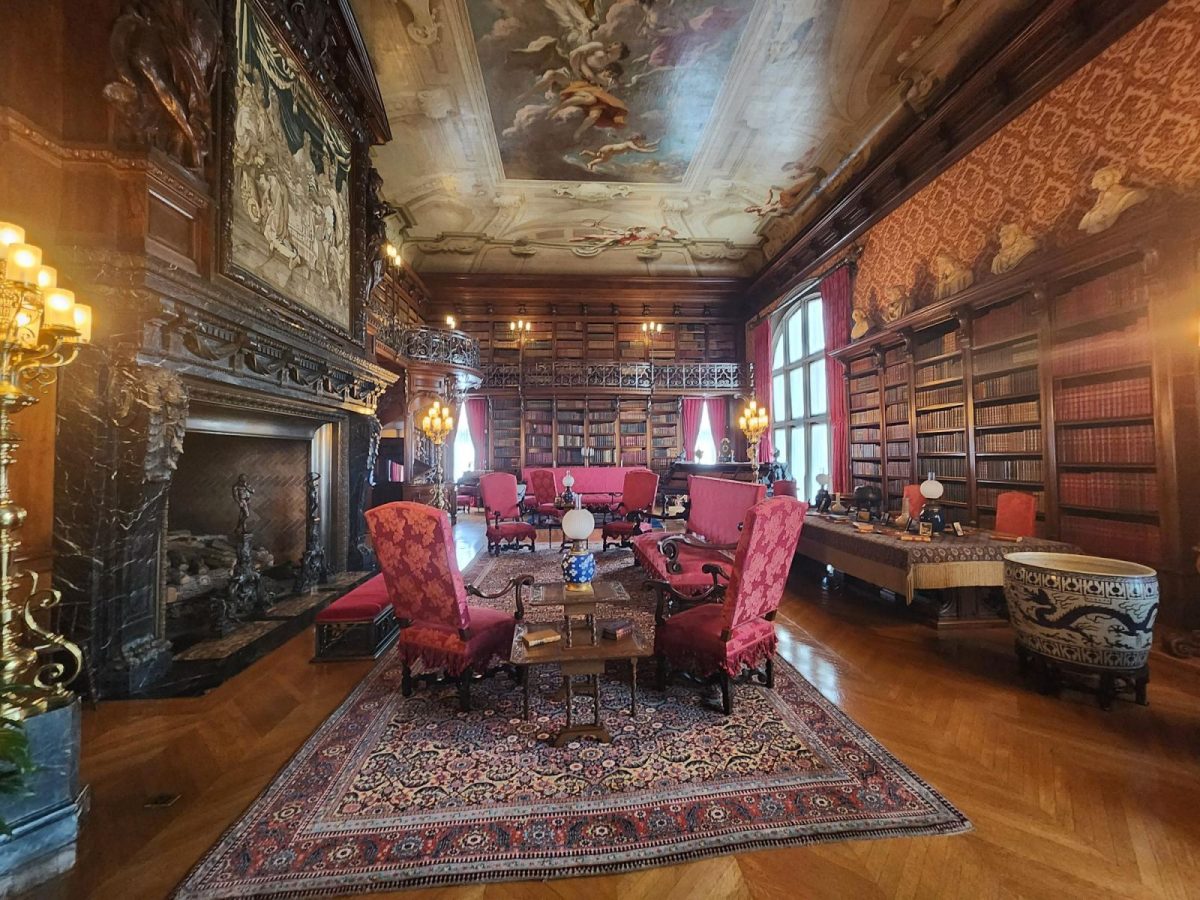 The Library inside Biltmore House, featuring floor-to-ceiling bookshelves, red upholstered seating, an ornate fireplace, historic tapestries, and a painted ceiling.