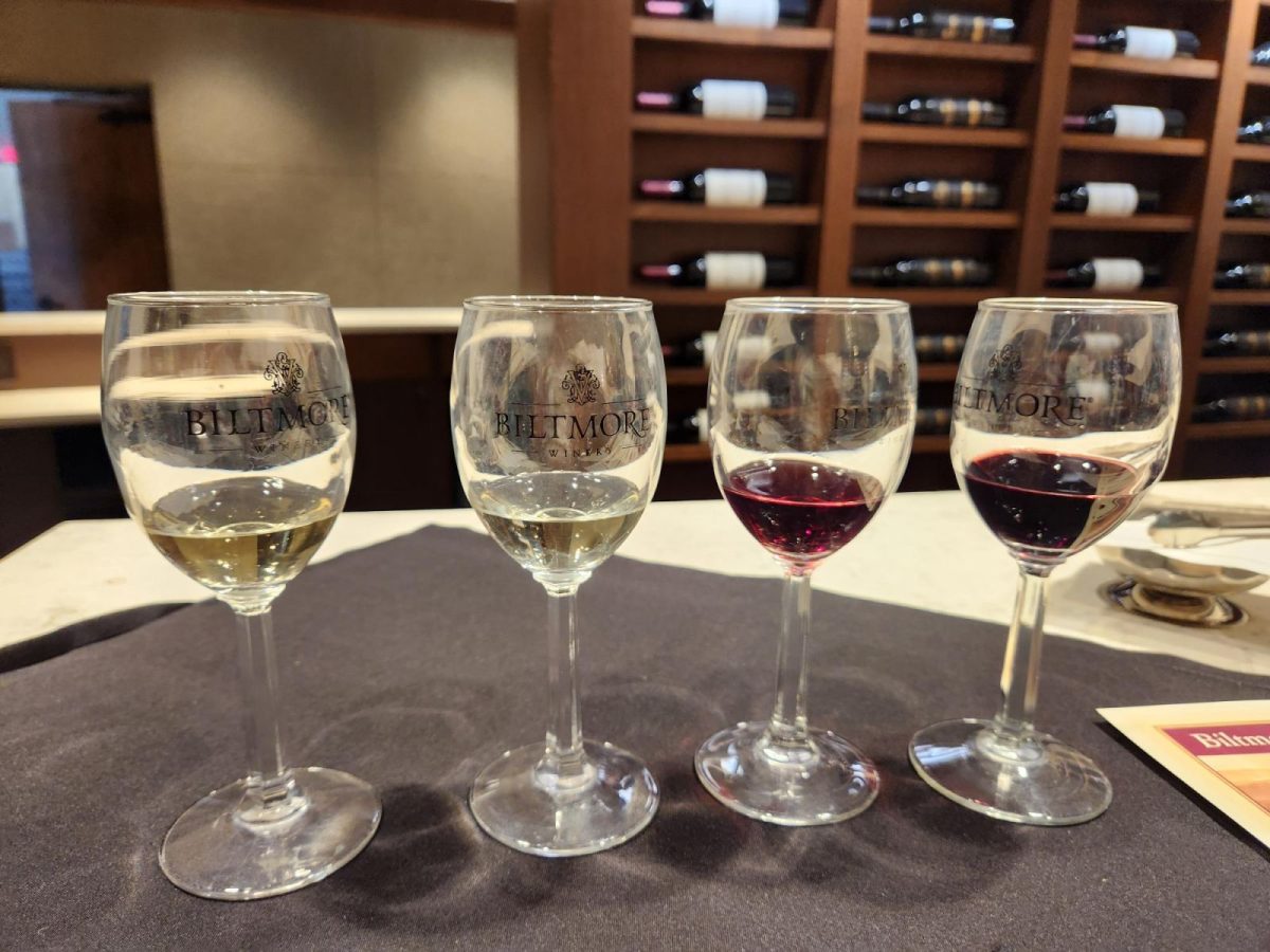 Four glasses of white and red wine arranged for a tasting at Biltmore Winery, with wine bottles displayed on wooden shelves in the background.