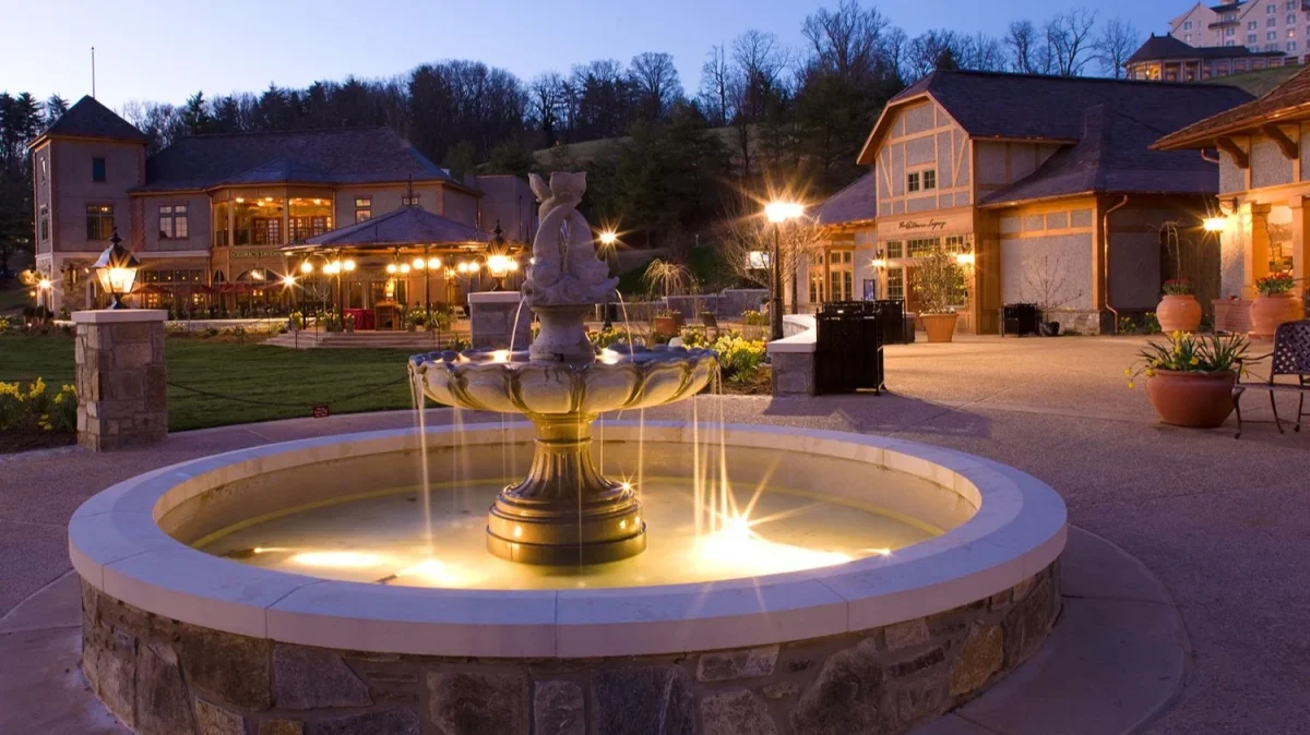 Illuminated fountain in the courtyard of Antler Hill Village at Biltmore Estate, surrounded by warmly lit buildings, gardens, and outdoor gathering spaces at dusk.