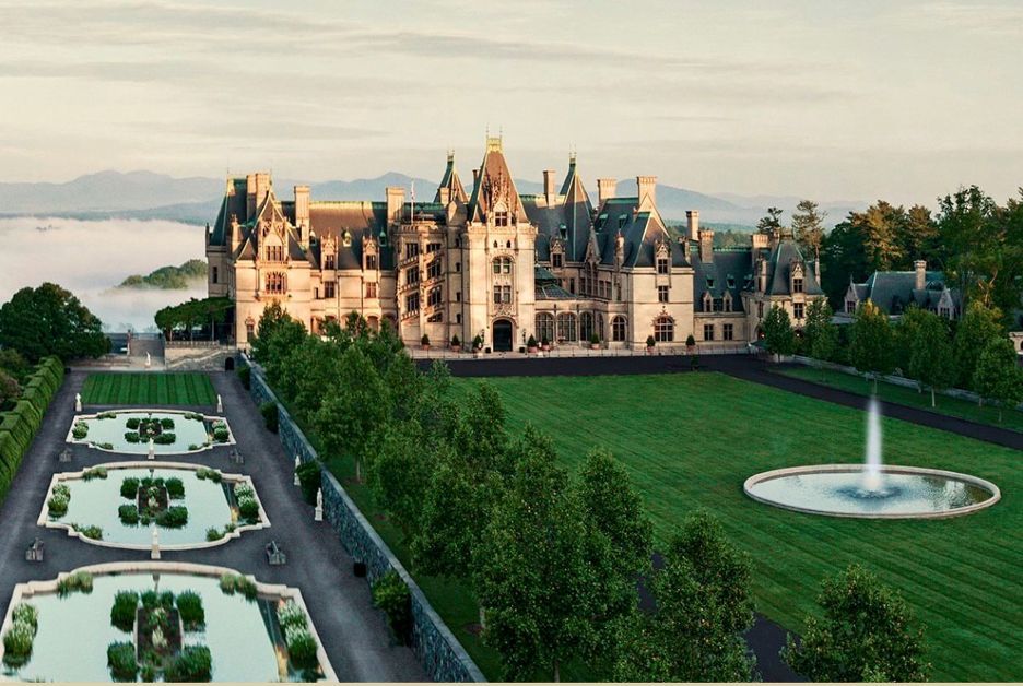 A wide view of Biltmore Estate in Asheville, North Carolina, showcasing the historic French Renaissance château overlooking its formal gardens, reflecting pools, expansive front lawn, and surrounding Blue Ridge Mountains at sunrise.