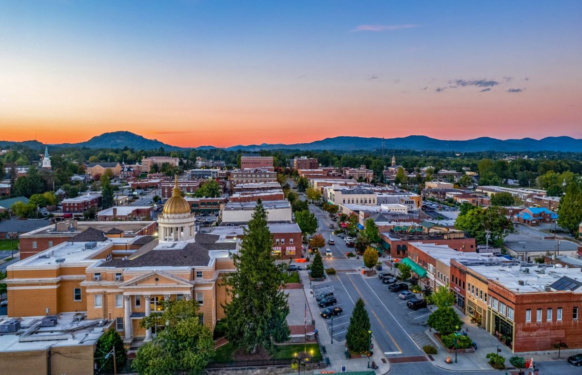 Aerial view of downtown Hendersonville, North Carolina, with the historic courthouse, Main Street buildings, and Blue Ridge Mountains beneath a colorful sunset sky.
