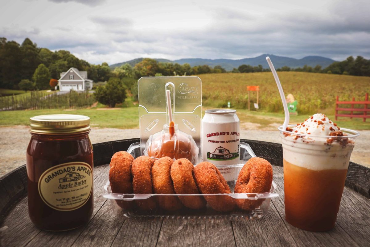 Apple butter, fresh apple cider, apple cider doughnuts, and a whipped cider drink from Grandad’s Apples displayed on a wooden table overlooking an orchard and the Blue Ridge Mountains.
