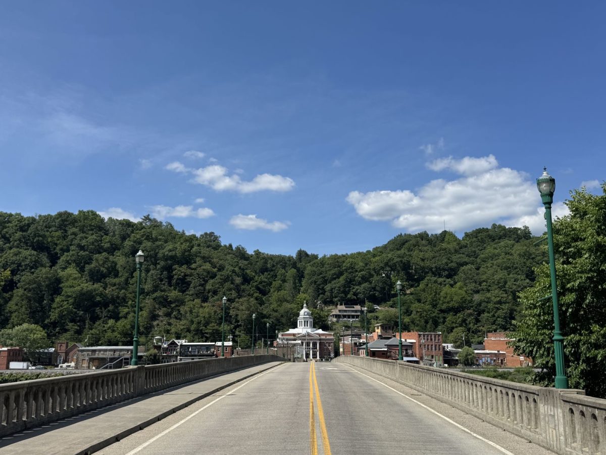 A view from the bridge leading into downtown Marshall, North Carolina, with the historic Madison County Courthouse centered in the distance. The French Broad River valley is framed by lush, forested mountains under a bright blue summer sky with scattered white clouds.