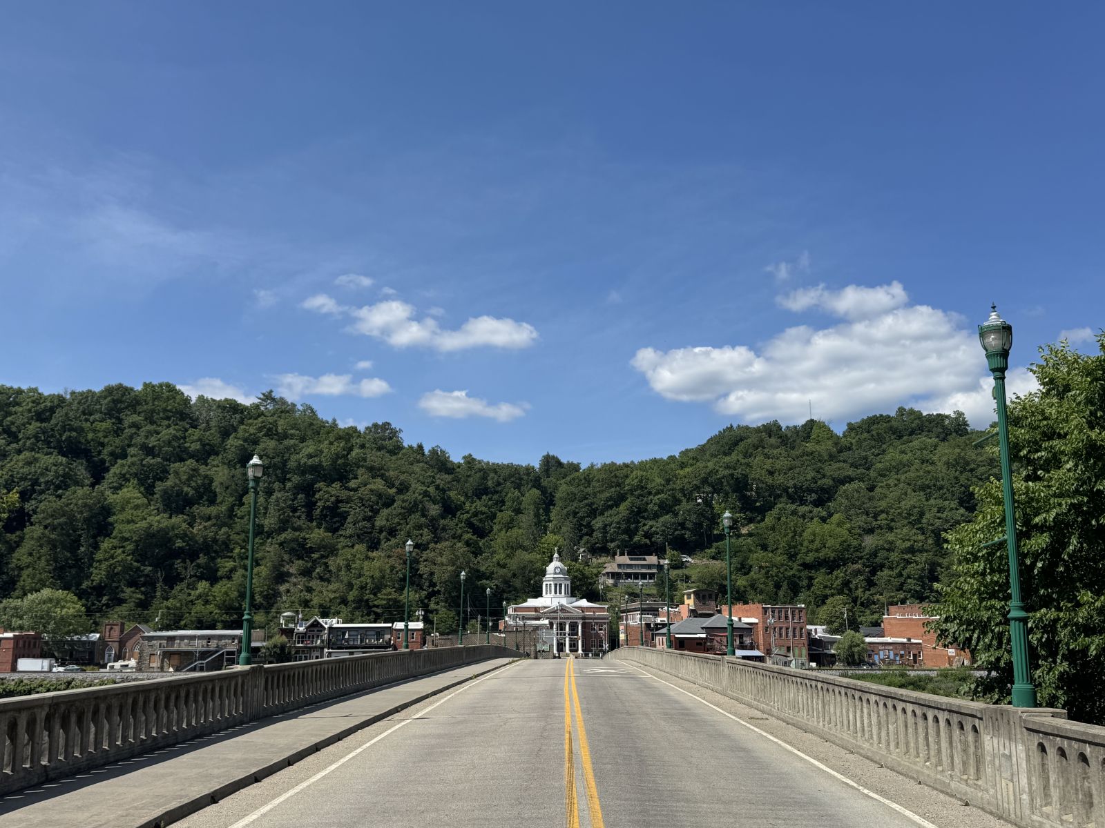 Bridge stretches toward a hillside town with a white-domed building, flanked by green hills and decorative street lamps under a blue sky.