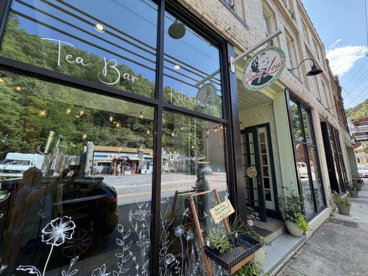 The storefront of Ginny Lou's in downtown Marshall, North Carolina, featuring large front windows, a hanging shop sign, and hand-lettered window graphics highlighting its tea bar and curated kitchenware.