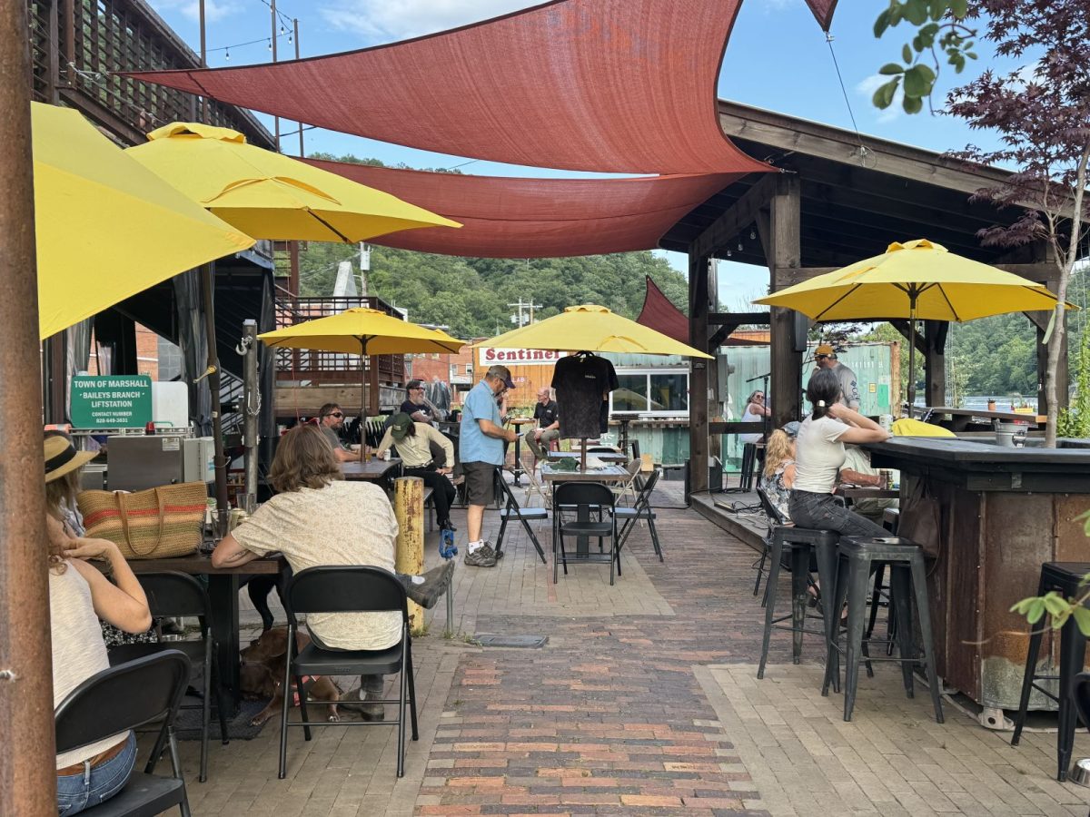 Outdoor patio in downtown Marshall, North Carolina, with colorful shade sails, bright yellow umbrellas, and guests enjoying drinks and conversation in a lively gathering space.