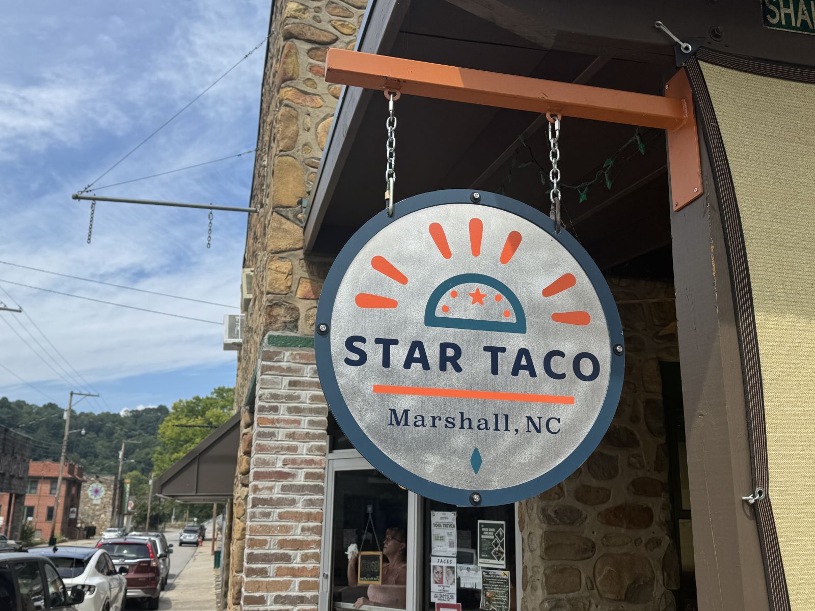 Round Star Taco hanging sign outside the restaurant in downtown Marshall, North Carolina, featuring the restaurant's logo against a backdrop of historic buildings, blue sky, and mountain scenery.