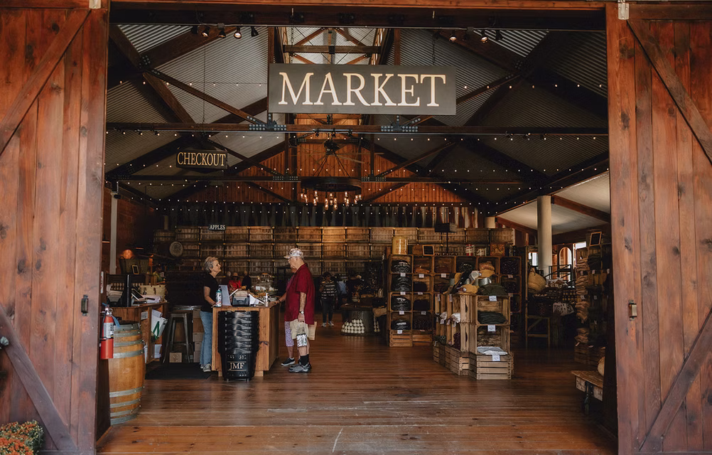 Open entrance to a rustic farm market with wooden floors, exposed beams, string lights, produce displays, and shoppers near the checkout counter.