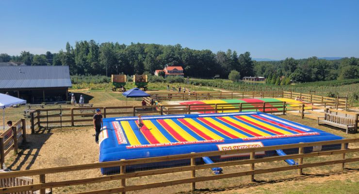 Children play on a large colorful jumping pillow at a family-friendly apple orchard surrounded by farm fields and mountain scenery.