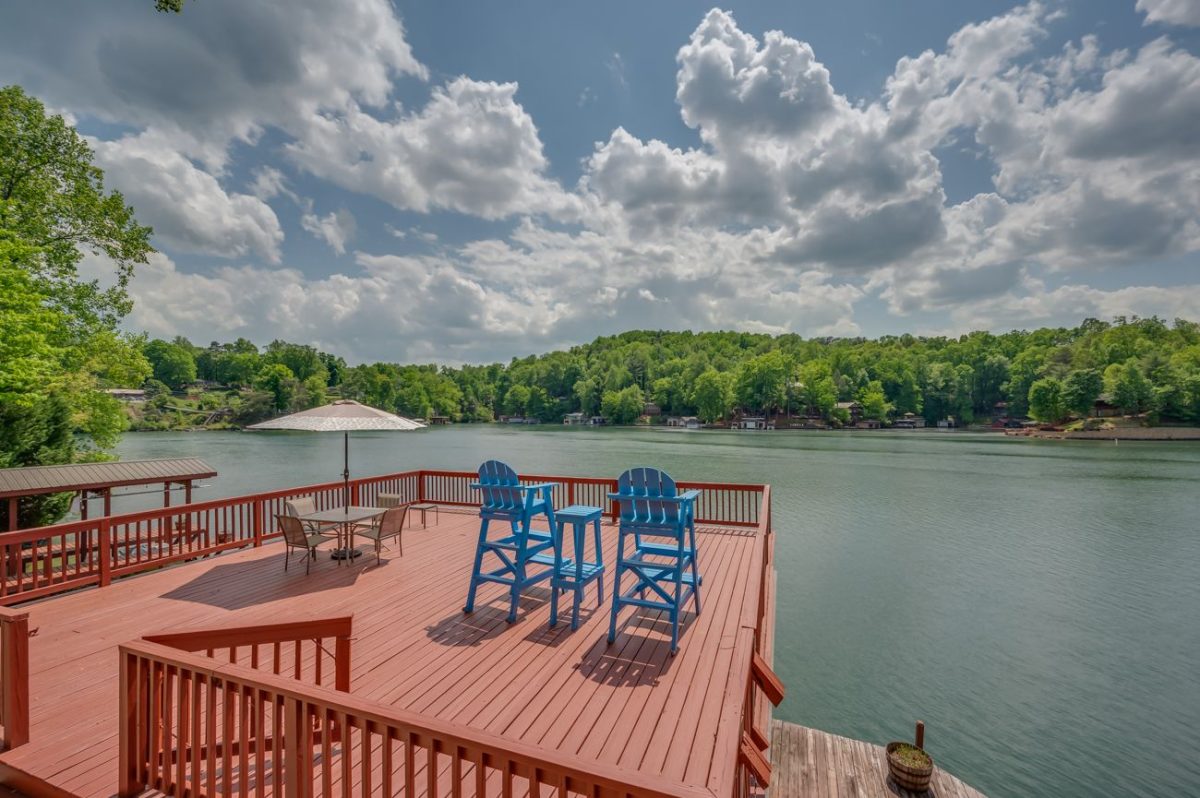 Expansive lakeside deck at M&M Rockhouse on Lake Lure featuring bright blue Adirondack chairs, outdoor dining, a private dock, and panoramic water views surrounded by lush Blue Ridge Mountains.