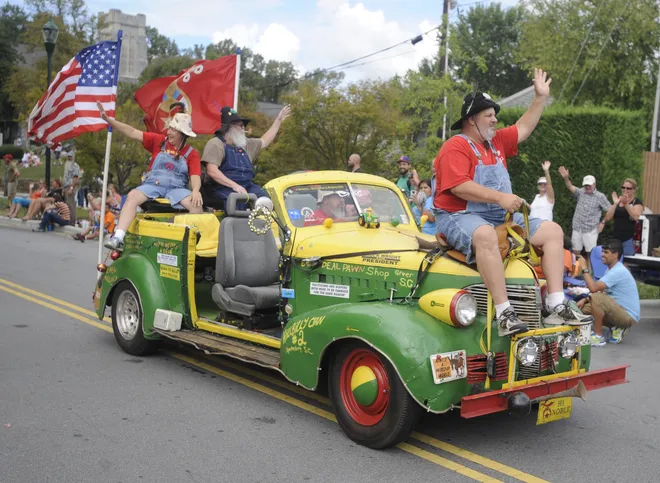 Three men in overalls and hats wave from a colorful vintage pickup truck decorated for a community parade as spectators watch from the roadside.