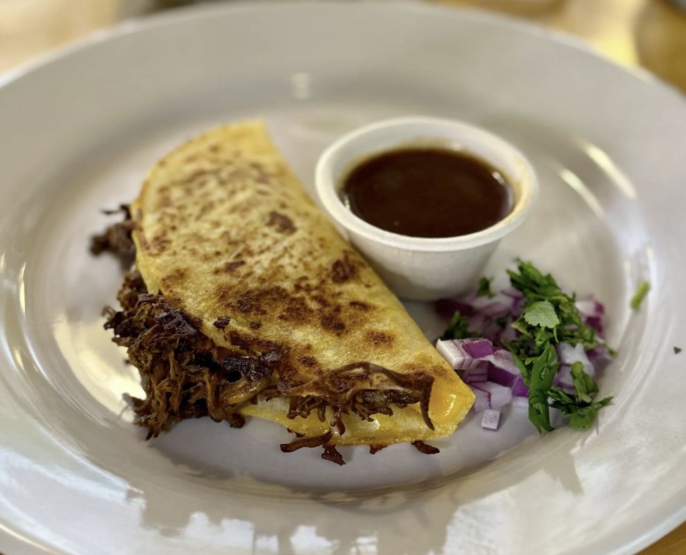 A golden-brown birria taco from Star Taco, filled with slow-cooked shredded beef and melted cheese, served with a side of rich consommé, diced red onions, and fresh cilantro on a white plate.