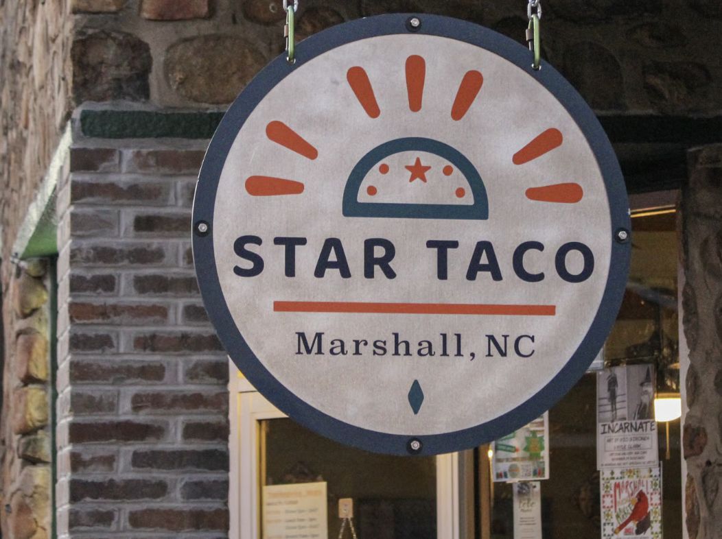 The round hanging sign for Star Taco in downtown Marshall, North Carolina, displayed outside the restaurant against a backdrop of brick and stone, featuring a stylized taco logo and the words "Star Taco – Marshall, NC."