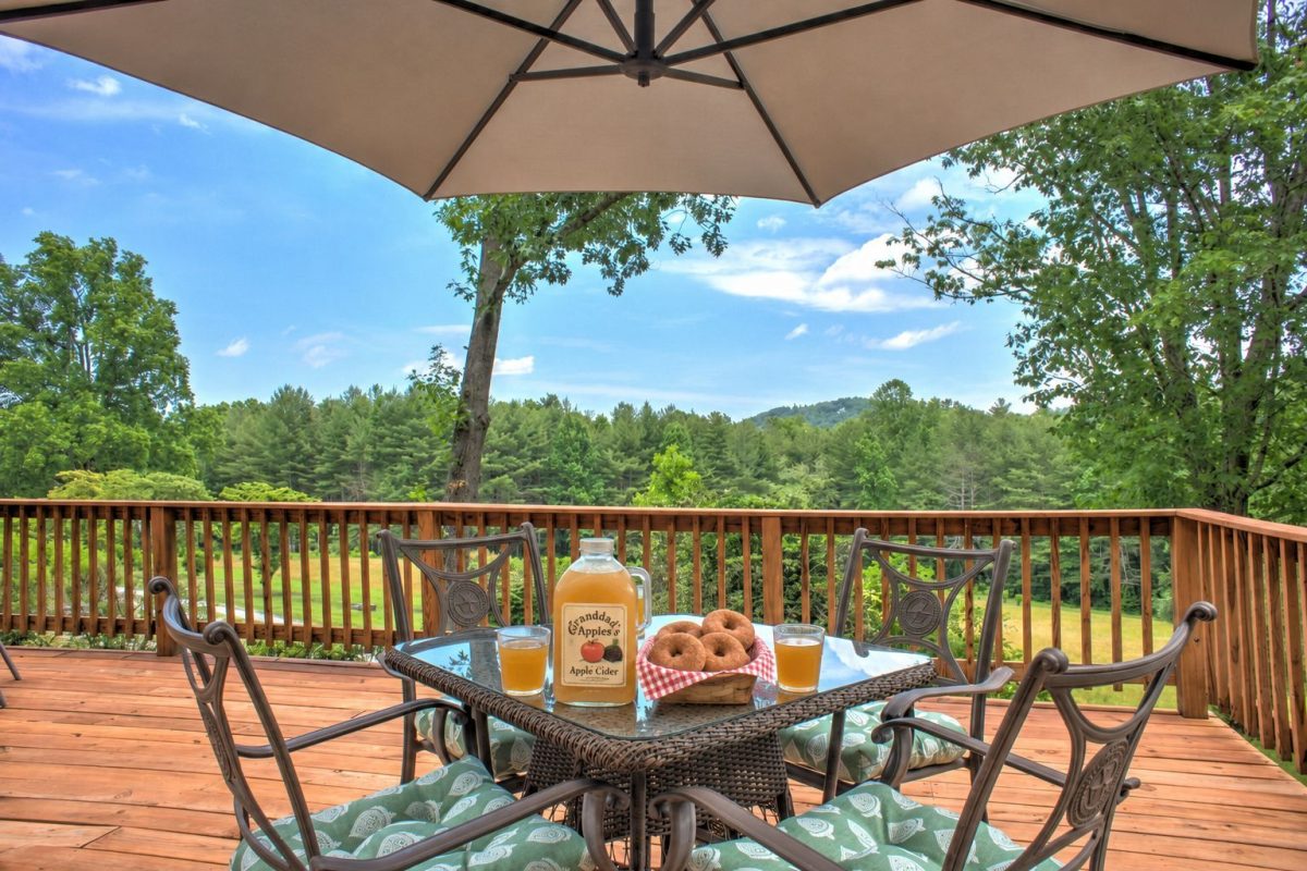 Outdoor deck overlooking a wooded mountain landscape, with Grandad’s Apples cider, two glasses of cider, and a basket of apple cider donuts arranged on the patio table.