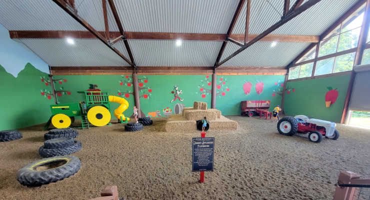 Indoor farm-themed play area with child-sized tractors, stacked hay bales, large tires, a tractor-shaped slide, and colorful orchard murals.