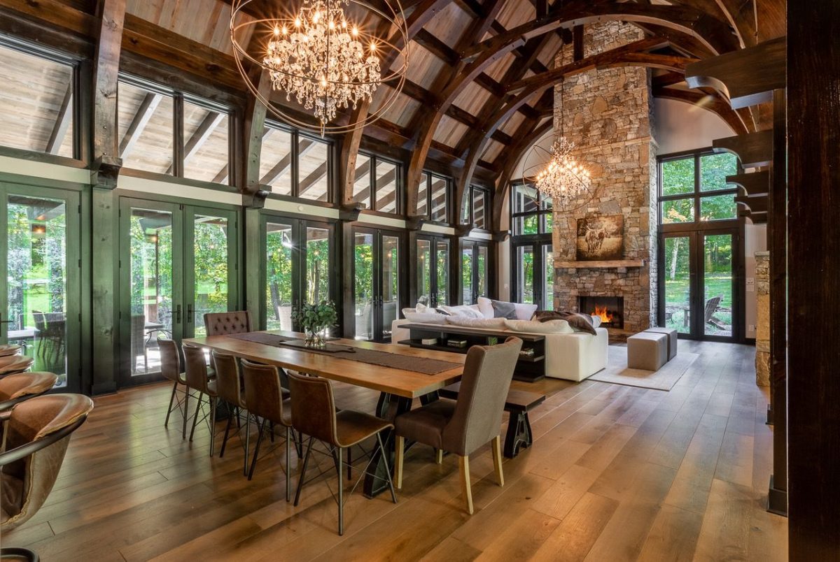 Rustic modern living and dining area with a long wooden table, leather chairs, and a stone fireplace under a vaulted wooden ceiling with chandeliers.