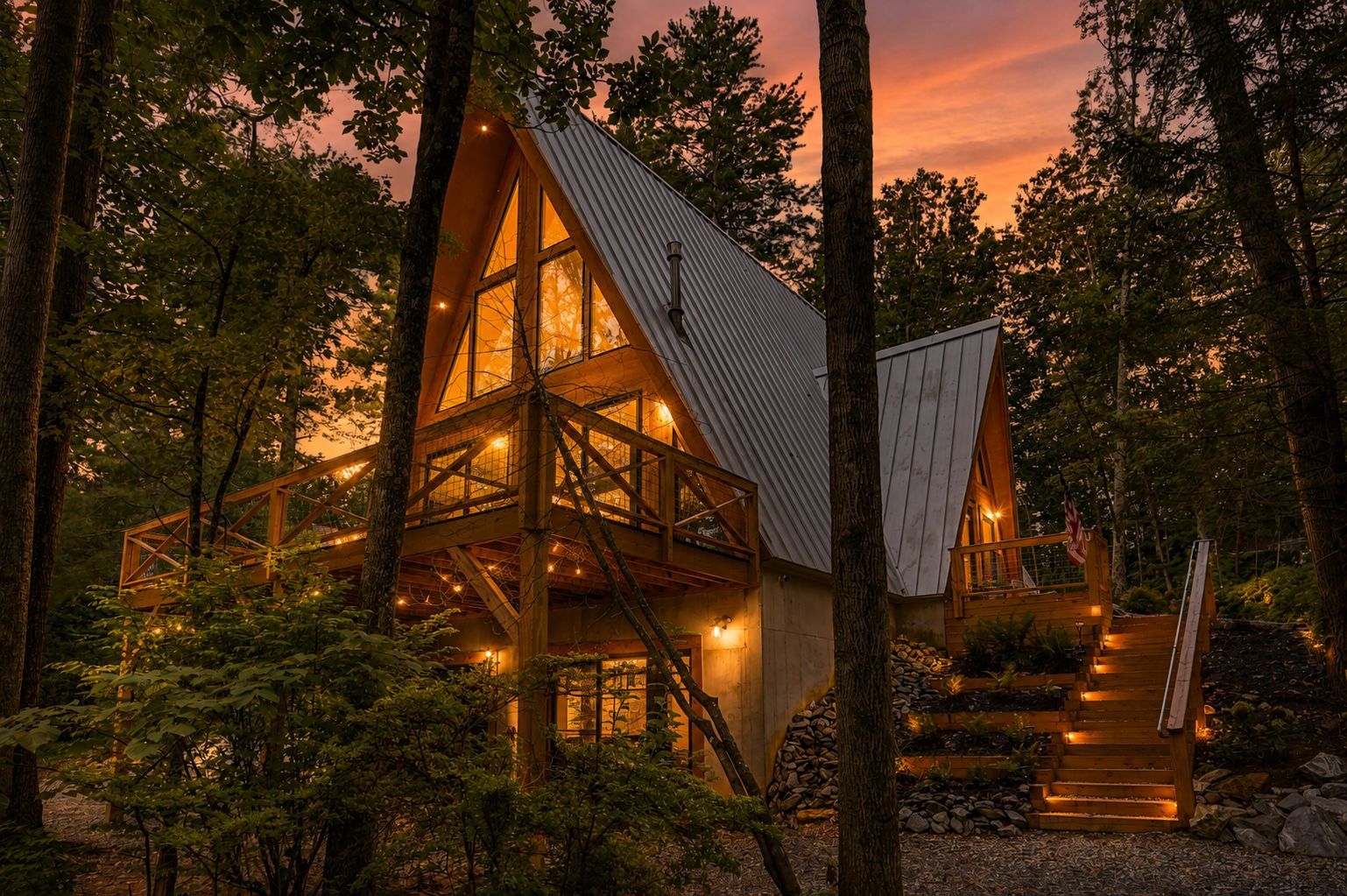 Twilight view of the A-Frame Nest, a modern A-frame cabin in Asheville, North Carolina, glowing warmly beneath a colorful sunset sky and surrounded by a peaceful forest.