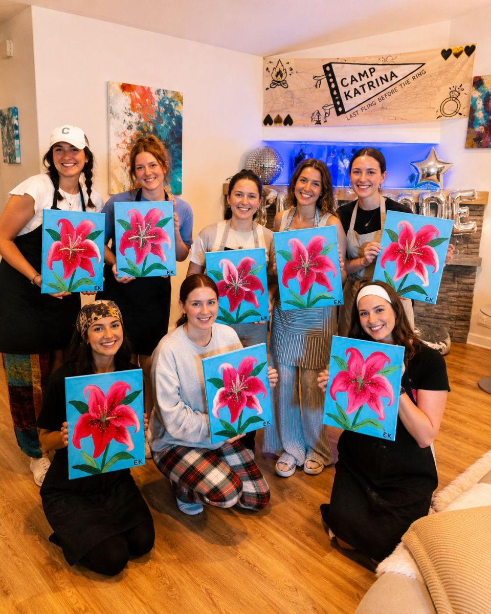 Nine people in a cozy studio hold blue canvases with pink lilies, smiling at the camera. A Camp Katrina banner hangs in the background.