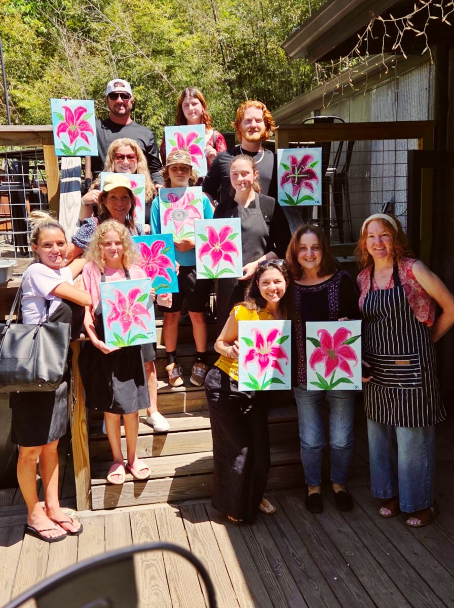 Group of about 12 people on a deck, proudly displaying bright pink lily paintings they've created outdoors.