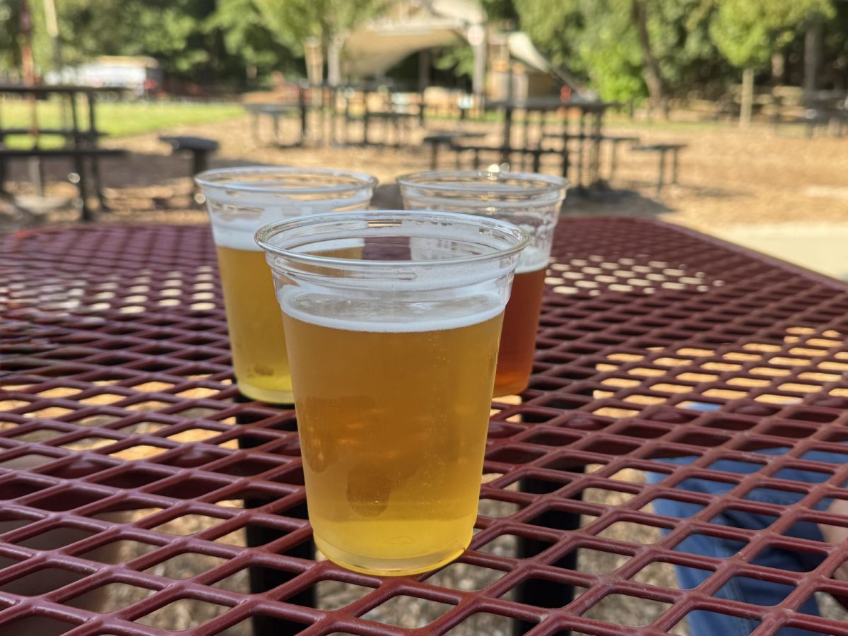 Three craft beers in clear tasting cups sit on a red picnic table at an outdoor brewery beer garden, with picnic seating and a shaded stage softly blurred in the background.