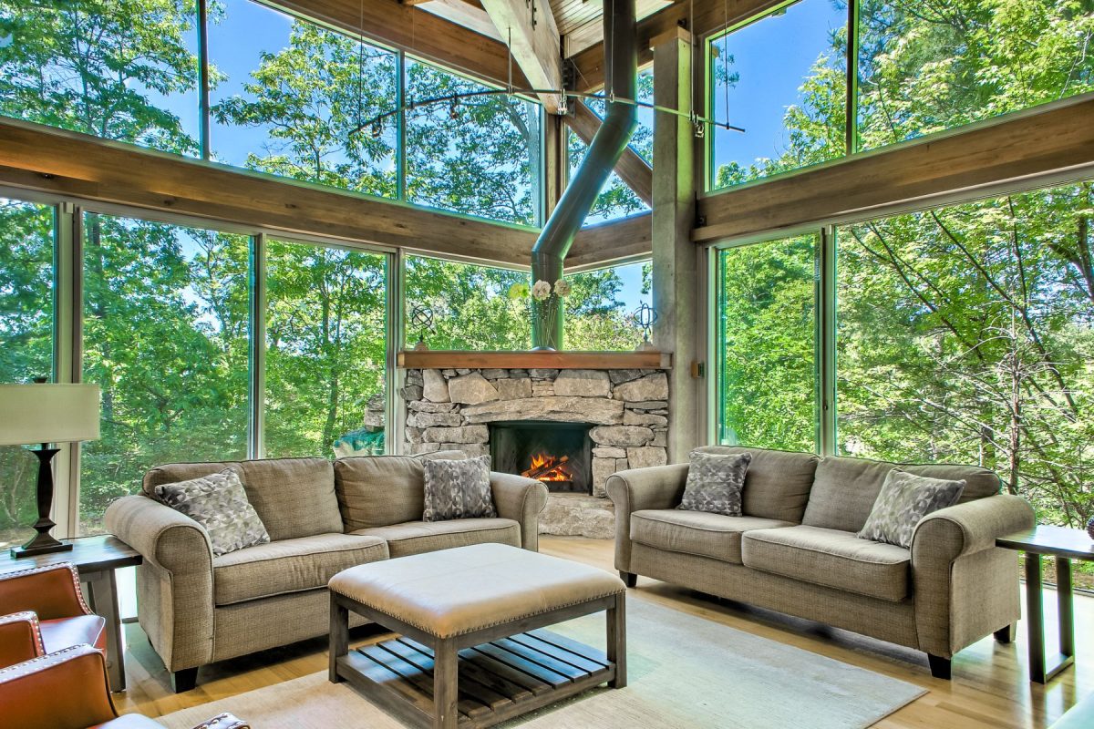 Light-filled great room at Innisfree Farms featuring floor-to-ceiling windows, a stone fireplace, vaulted wood-beam ceilings, and comfortable seating surrounded by peaceful forest views.