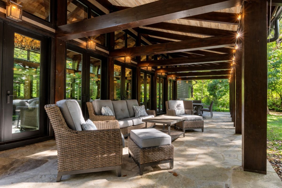 Covered outdoor patio at Modern Mountain Creekhouse featuring comfortable wicker lounge seating, a stone floor, exposed timber beams, string lights, and peaceful wooded surroundings in Asheville, North Carolina.