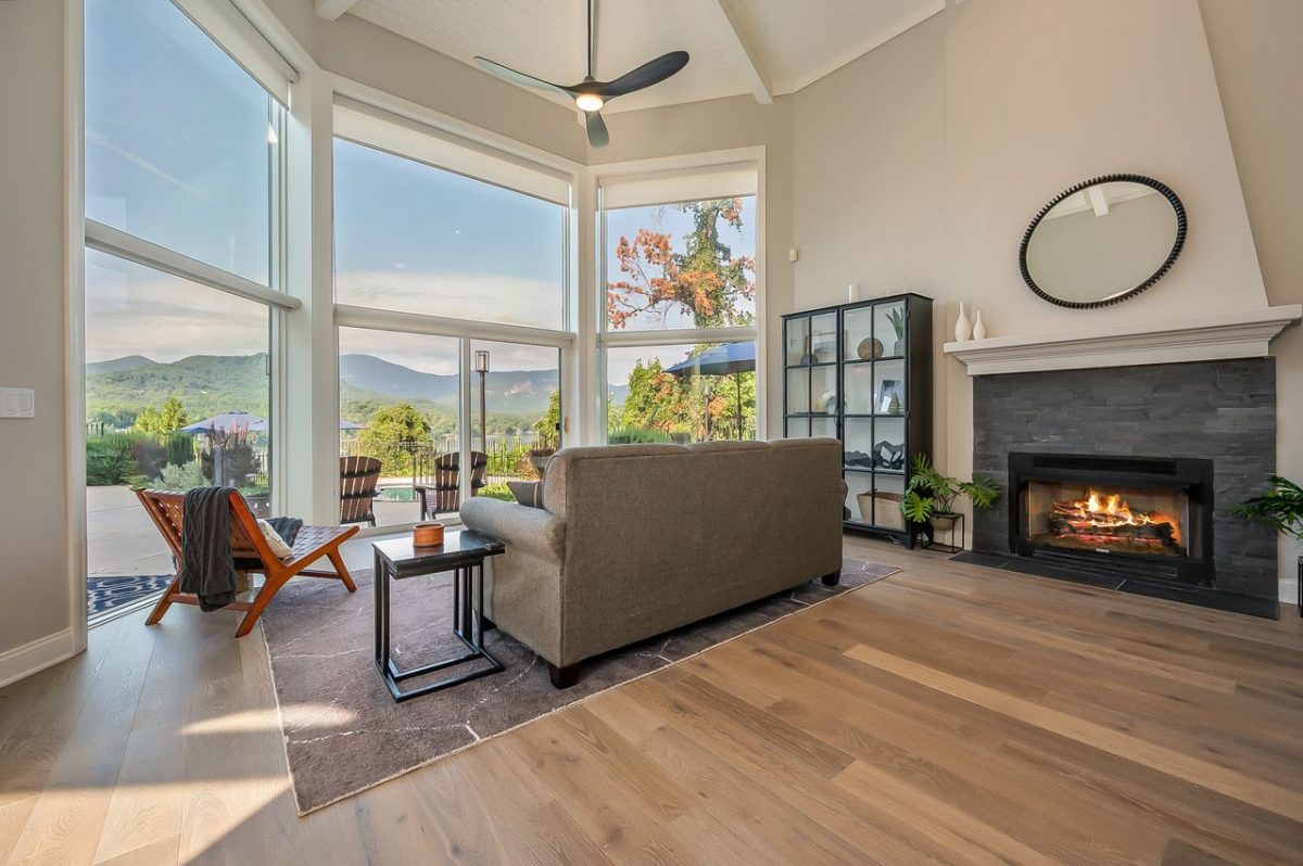 Bright living room with large windows overlooking mountains, a gray sofa, and a stone fireplace with a mantel.