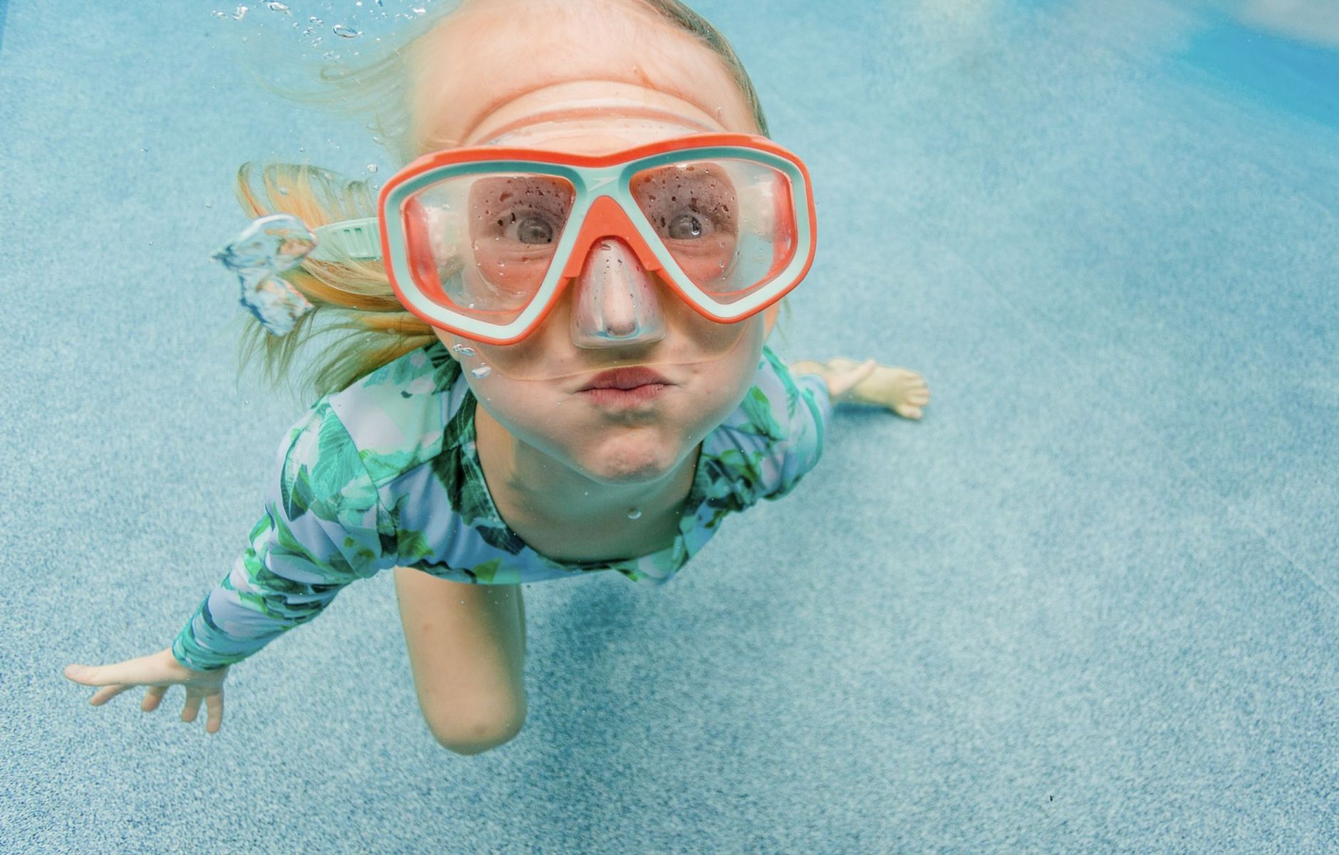 Young child wearing a bright snorkel mask swims underwater in a clear swimming pool, looking directly at the camera with bubbles rising around them during a summer vacation.