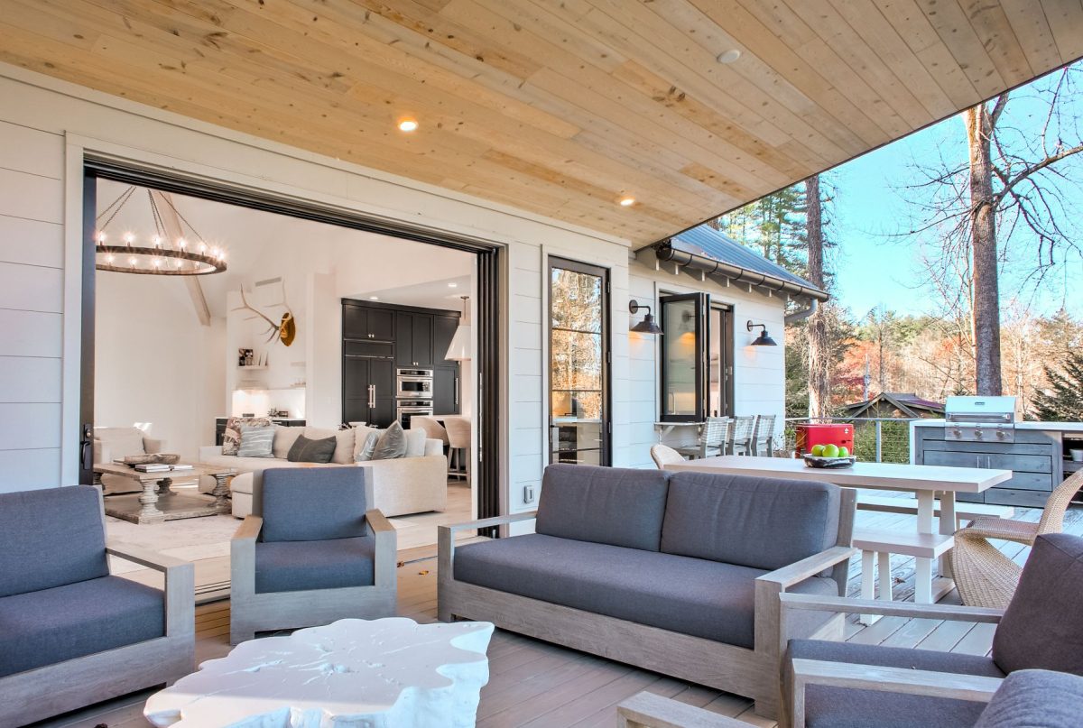Covered outdoor living space at The Summer Camp featuring comfortable lounge seating, a dining table, built-in grill, and wide folding glass doors that open to a bright, modern great room. The seamless indoor-outdoor design creates an inviting space for relaxing and entertaining in the Blue Ridge Mountains.
