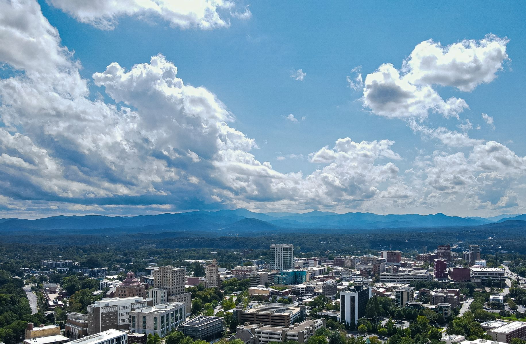 Aerial view of downtown Asheville, North Carolina, with the Blue Ridge Mountains stretching across the horizon beneath dramatic summer clouds and blue skies.