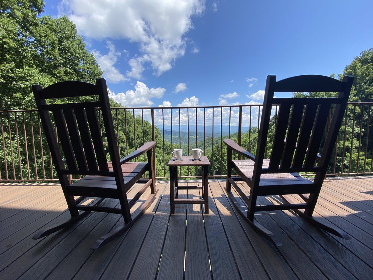 Two wooden rocking chairs and morning coffee on a mountain-view deck overlooking the rolling Blue Ridge Mountains beneath a bright blue summer sky.